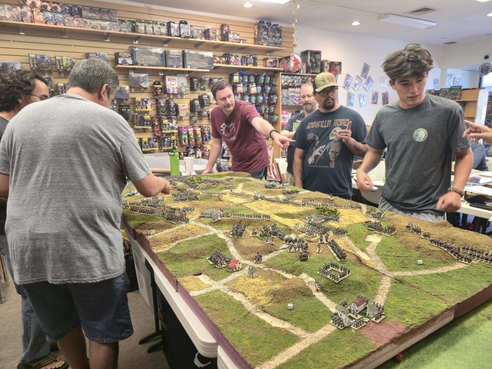 A number of men surround a large wargames table, with small painted miniatures visible, and game store merchandise in the background.