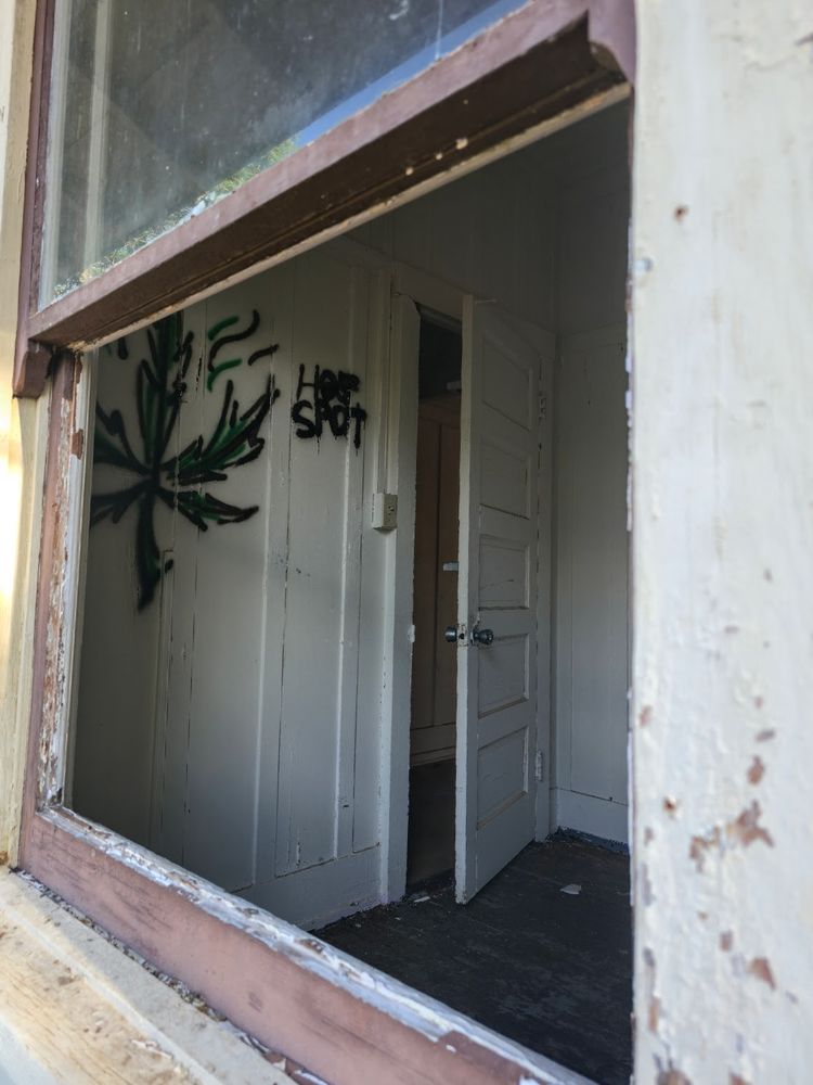 View through a broken window into a small, empty room with peeling paint and dark wooden floors. Inside, graffiti of a marijuana leaf and the words 