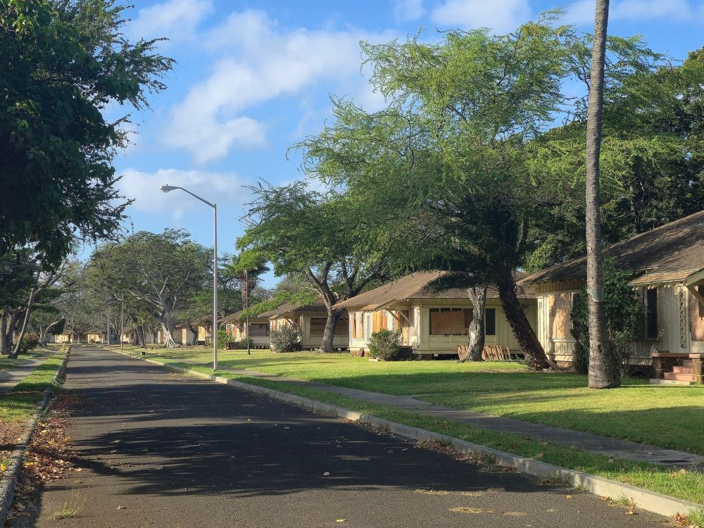 A quiet, sunlit, empty street at Ft. Kamehameha lined with boarded-up, single-story wooden houses. The homes are spaced evenly along a grassy strip with trees, providing partial shade. The road is empty, with drifts of leaves against one curb, and a streetlamp stands on the sidewalk. The overall atmosphere suggests abandonment or disuse.