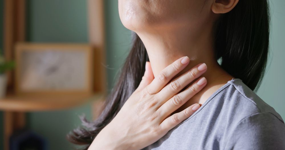 Close up of a woman's lower face and neck, she has her hand over her neck feeling it. She's wearing a blue t-shirt and has shoulder length black hair. 