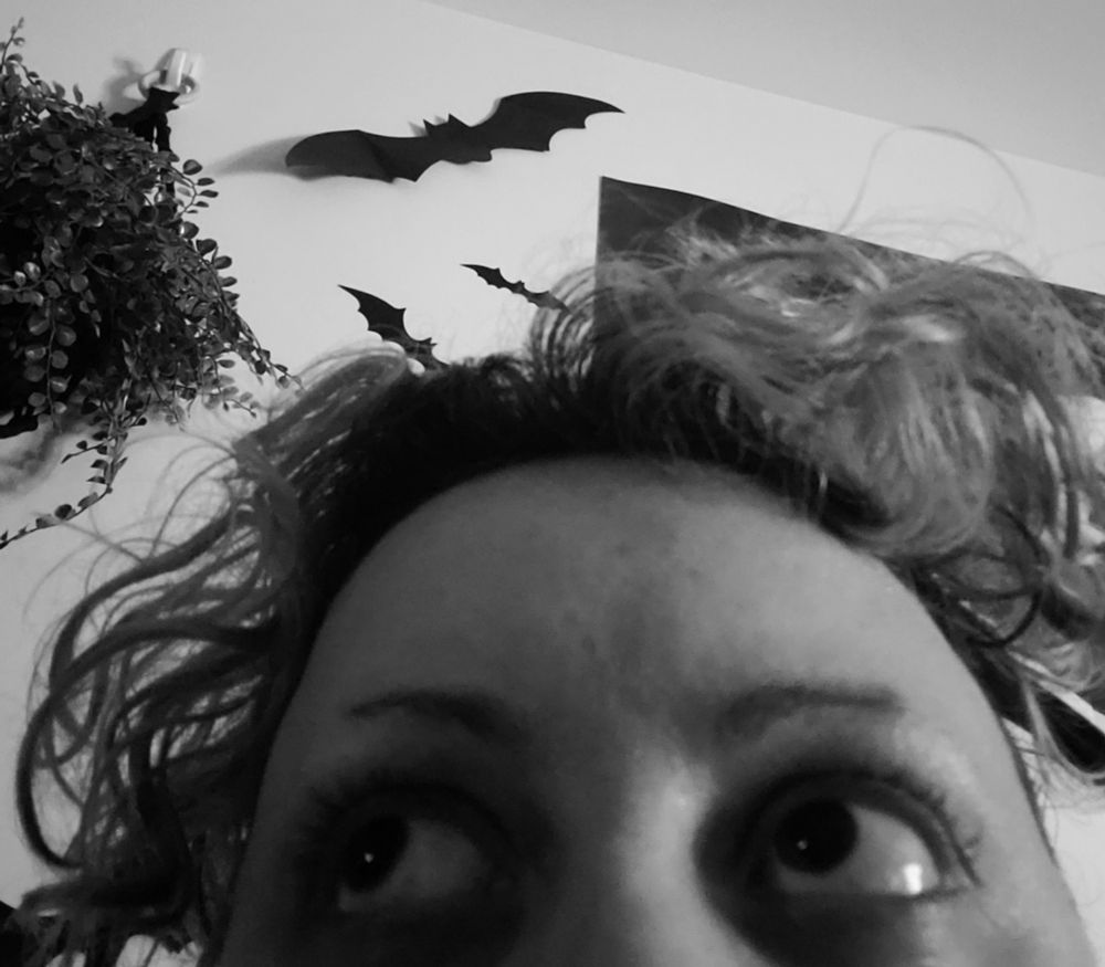 A black, grey & white toned photo of me, a non-binary person with large round eyes & short loose curly hair, looking up to the side. Background has a large vintage movie poster of Frankenstein's Bride, a few fake bats surrounding it and a hanging plant.