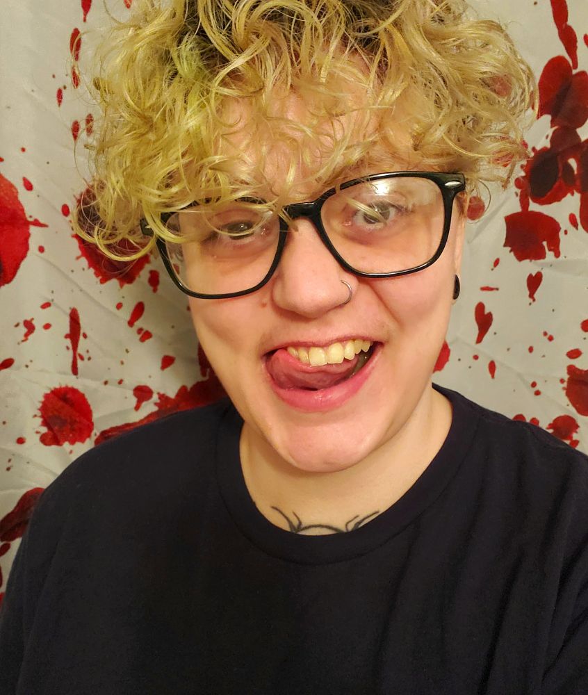A masculine leaning Non-Binary person with large black framed glasses, poofy curly bleached hair that is covering the forehead down to just mid ear. They're making a cheeky smile with their tongue up touching part or their smile. The background is of a fake blood spattered shower curtain.