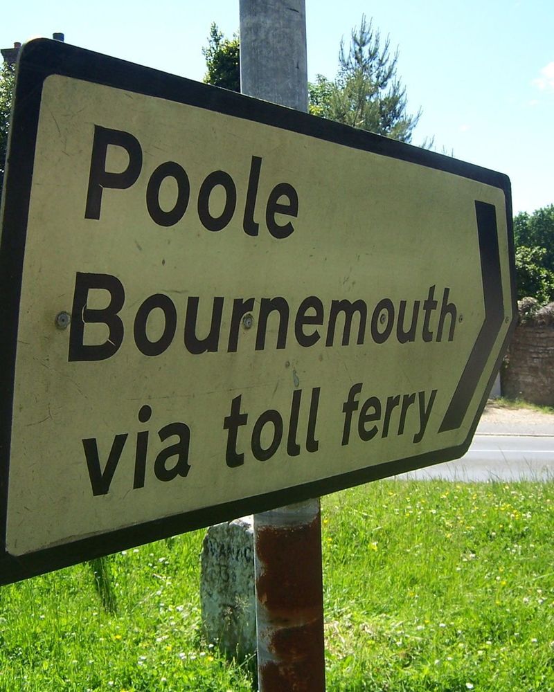 Photograph of an old direction sign pointing to the right, bearing the text "Poole, Bournemouth via toll ferry"