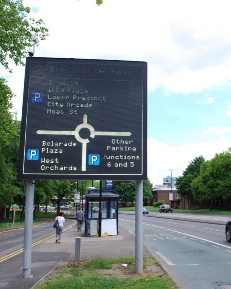 A huge electronic sign stands on the verge of an urban road, showing a diagram of a roundabout with car parks indicated on each of its three arms.