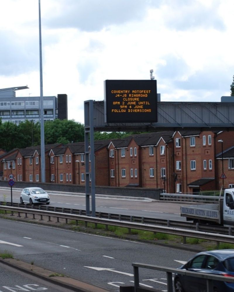 A busy urban dual carriageway passes in front of a row of houses. A metal gantry structure supports an electronic sign with a message about Coventry Motofest and road closure information.