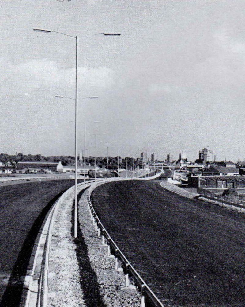Black and white photograph looking along an unfinished motorway. Two carriageways with a line of streetlights curve right then left into the distance between buildings