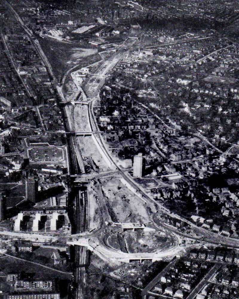 Black and white aerial photograph of an urban motorway under construction, with a route cleared through buildings and railway lines from a roundabout in the foreground away into the distance