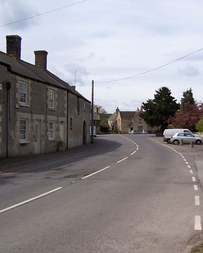 Photograph of a road passing through a quiet and picturesque village. A row of old stone cottages is on the left