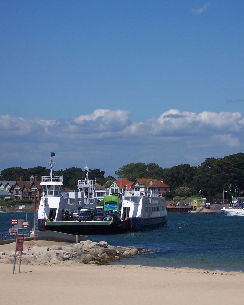 A small car ferry, painted white and loaded with vehicles, is seen mid-stream crossing a channel of water. A beach is in the foreground and the opposite shore is wooded with houses
