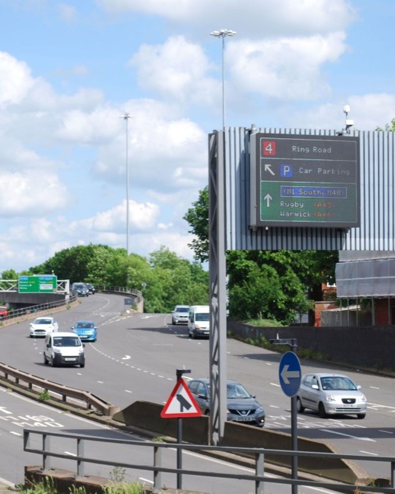 Photograph of an urban dual carriageway with traffic travelling and changing lanes as the road snakes towards the camera. An electronic sign is mounted on a gantry with the legend "Ring Road, Car Parking, (M1 South, M40), Rugby (A45), Warwick (A46)" shown in various colours.