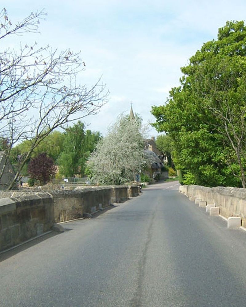 Photograph of a narrow road passing over a very long stone bridge with low walls on either side and trees overhanging in some places.
