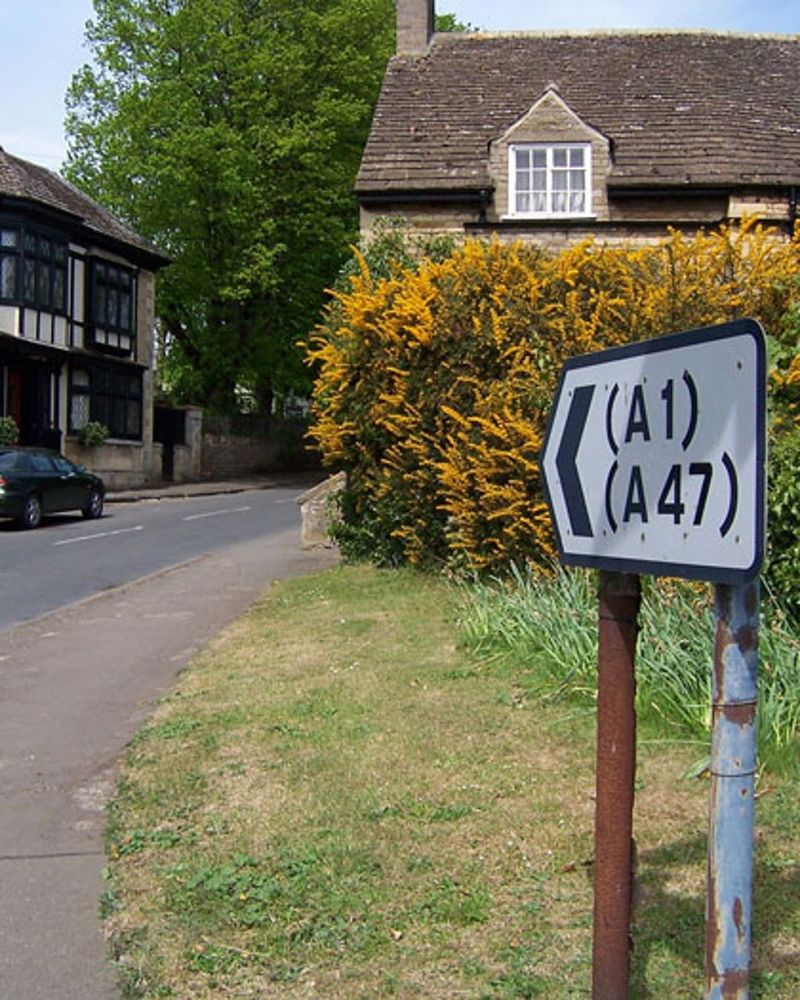 Photograph of a road in a quiet village, with a half-timbered house on the left and a stone cottage on the right obscured by hedges. In the foreground a direction sign points up the road, with the legend "(A1), (A47)"