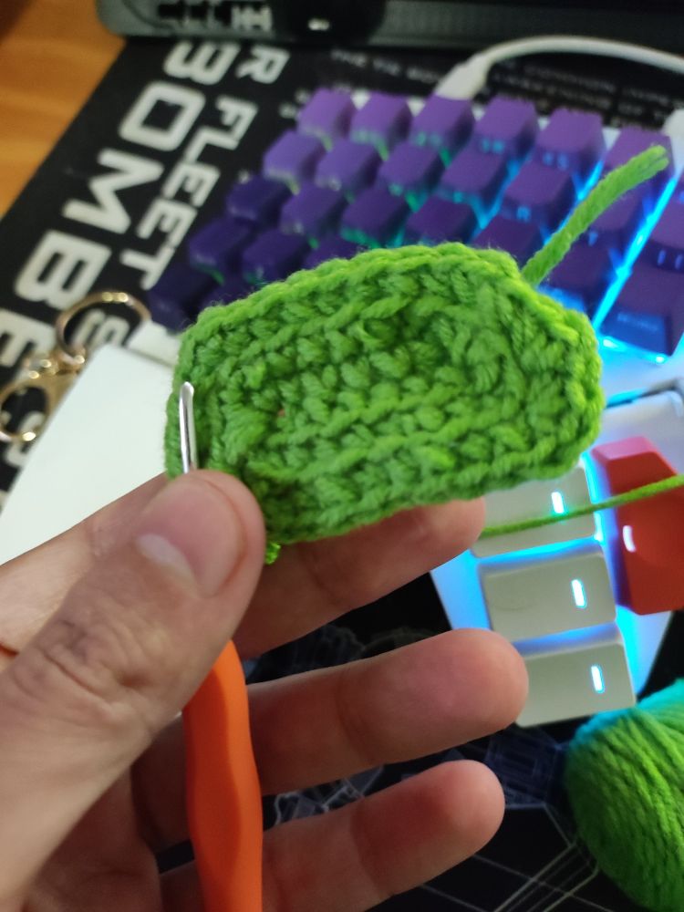 Uma mão segurando uma agulha e um quadradinho de crochet de barbante verde, ao fundo tem um teclado e um Mousepad 