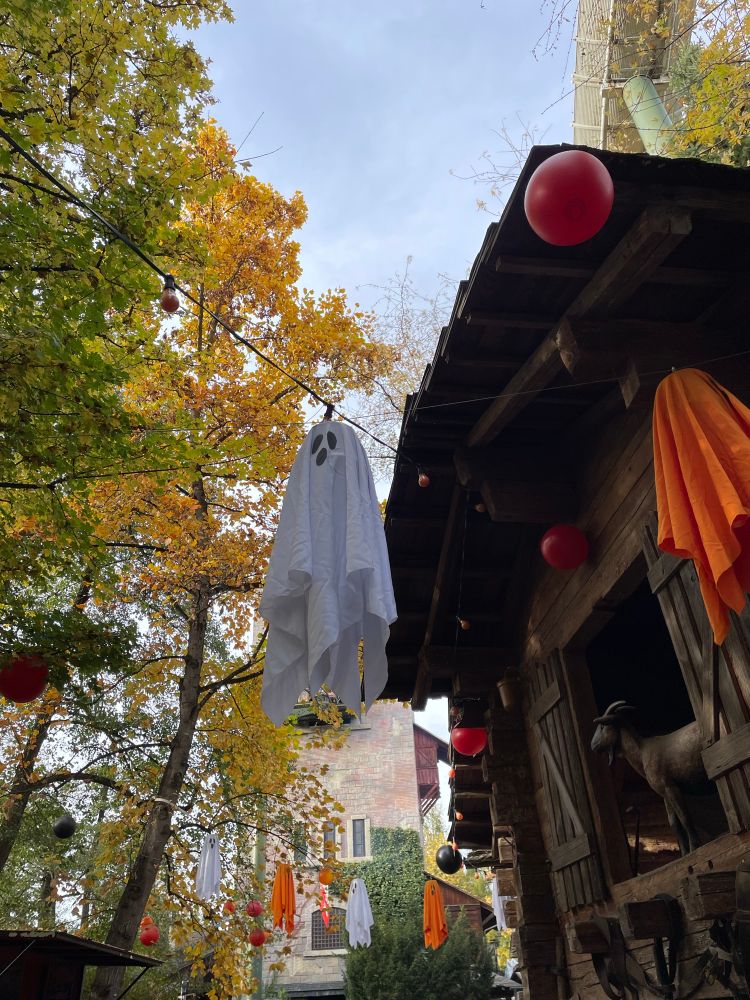 ein gespenst hängt an einer kette über einer straße, auf der rechten seite ein holzhaus, dahinter eine achterbahn 