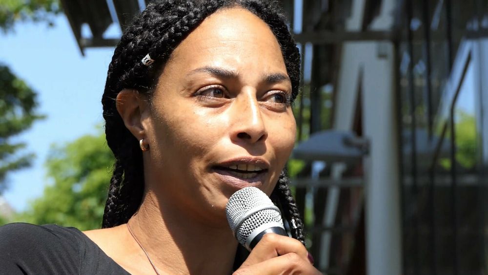 Een zwarte vrouw met braids die in een microfoon praat die ze zelf vasthoudt, met een onscherp gebouw en bomen op de achtergrond. 