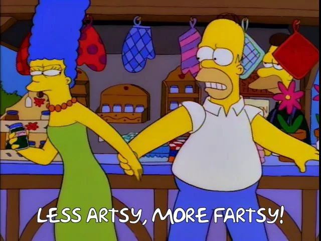 Still from The Simpsons at a chili cook off. Marge is trying to get Homer to look at the arts and crafts, and Homer wants to get to the beer and chili. Homer is saying “less artsy, more fartsy.”