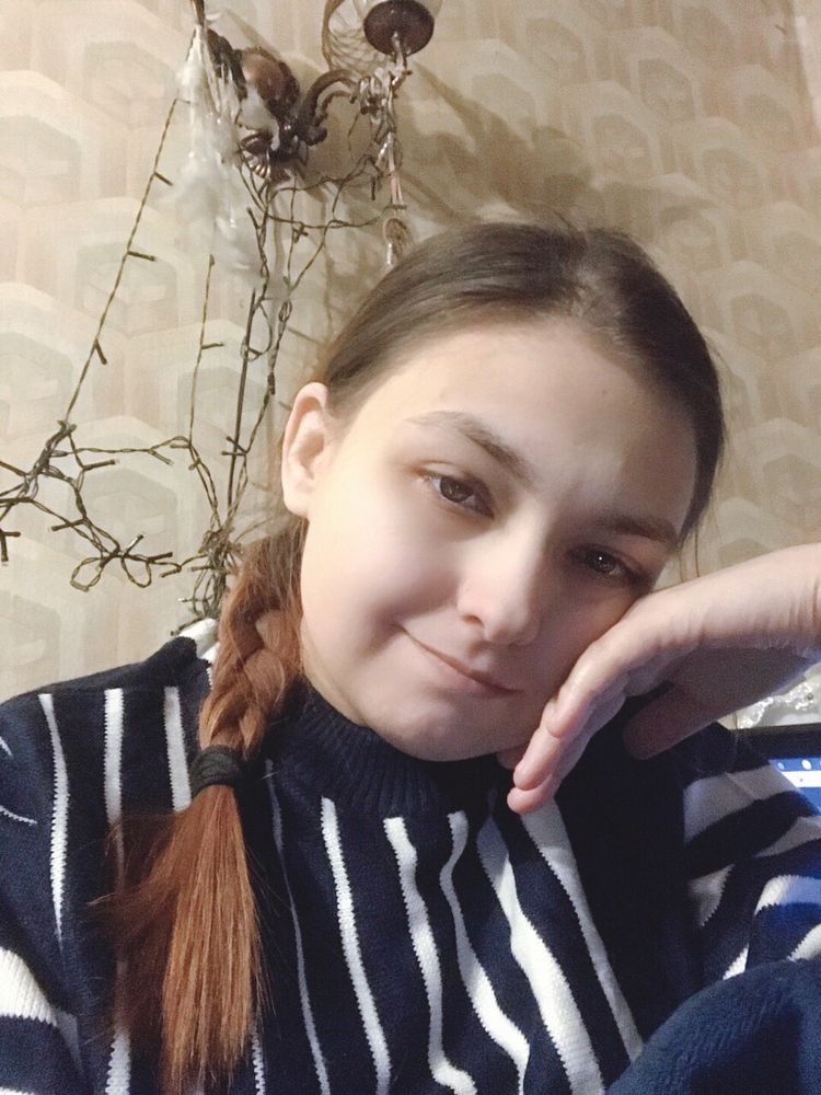 На фотографии на фоне бежевых обоев человек в свитере в сине-белую вертикальную полоску, с тёмными глазами и рыжеватыми тёмно-русыми волосами, заплетёнными в косичку, перекинутую на одно плечо.  Косичка перевязана чёрной резинкой.