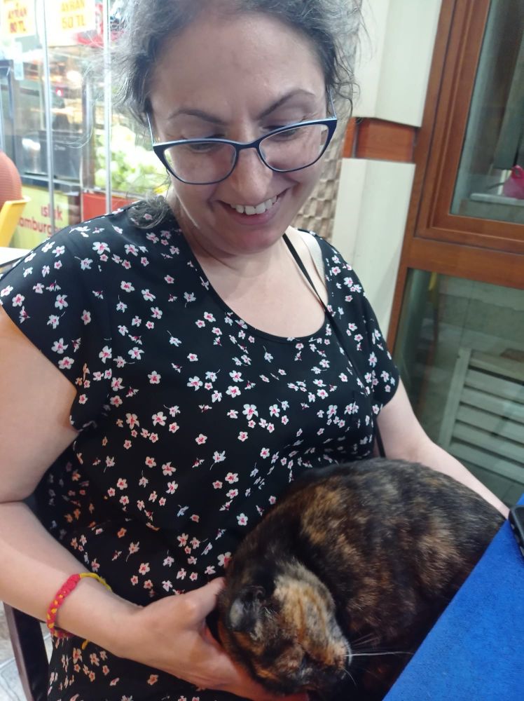 Me at a café in Türkiye with an adorable tortie cat curled up in my lap, purring like crazy. She protested when I had to stand up to leave. 
