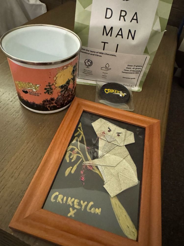 Swag given to Crikeycon X workshop creators: coffee beans, challenge coin, mug and frame of a dropbear origami.