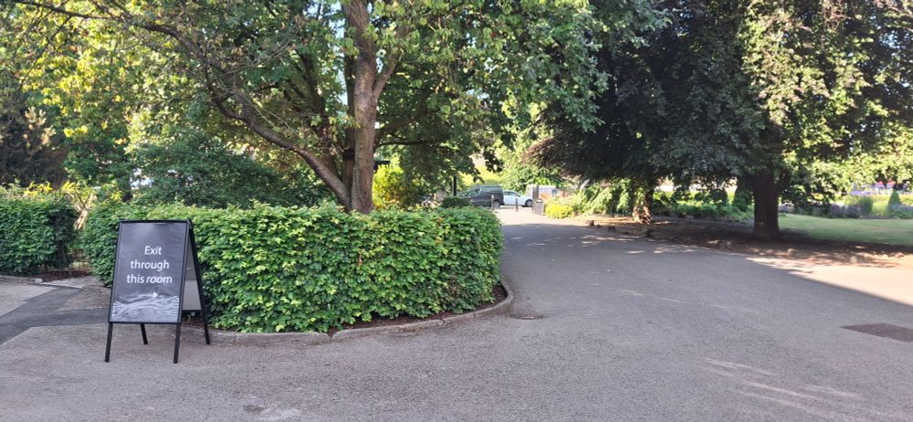 Photo shows a freestanding sign-board carrying a sign that says "Exit through this room", with an arrow pointing to the right. The sign is outside, with no building (and so no room) adjacent. To the right is a wide pathway surrounded by trees. 