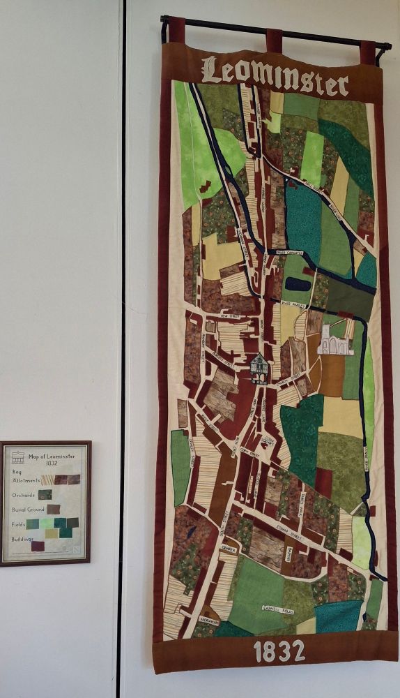 Photo of wall hanging showing a map of Leominster. Brown and green fabrics distinguish between fields, orchards and burial grounds. Roads and rivers are all labelled. The church and market hall are prominent buildings. A small frames piece hanging alongside provides the key to the map. 