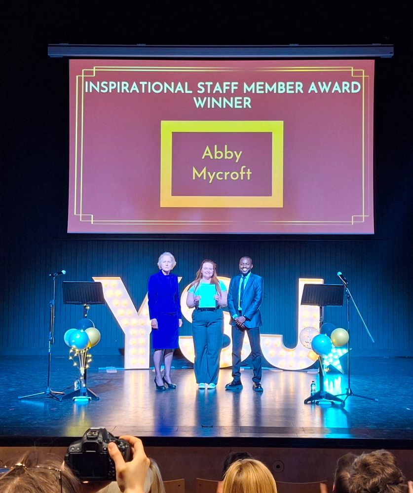 Abby on stage receiving her award from the Vice Chancellor and the SU President: Education. Behind them are the letter YSJ in lights. 