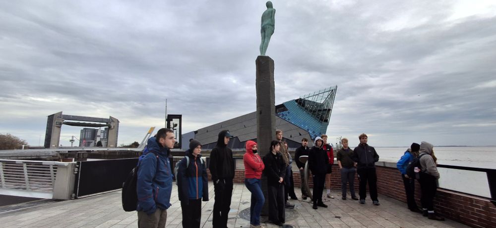 Students looking across the Humber Estuary, standing alongside the flood wall. The Deep aquarium is behind, and Voyage statue stands above them.