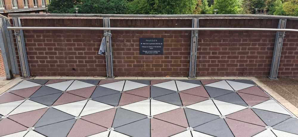 A grid of triangle tiles with sign on the wall behind explaining that walking across the tiles generates electricity, & inviting people to use the electricity to charge their phones on the benches nearby.  