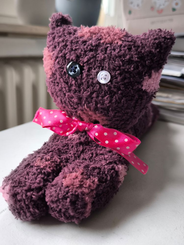 dark red sock poorly sewn into a cat, a black and a white button make up its eyes, a pink polka dot ribbon is tied into a bow around its neck