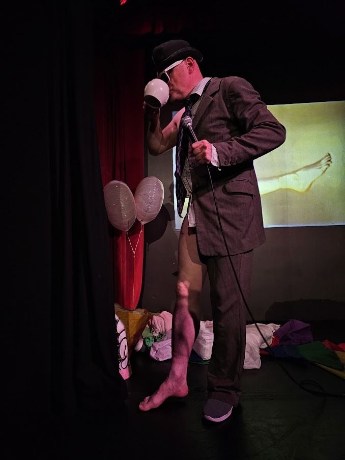 John Callaghan wearing half a suit divided lengthwise, sipping from a mug, with a projection of a sideways leg behind him. 