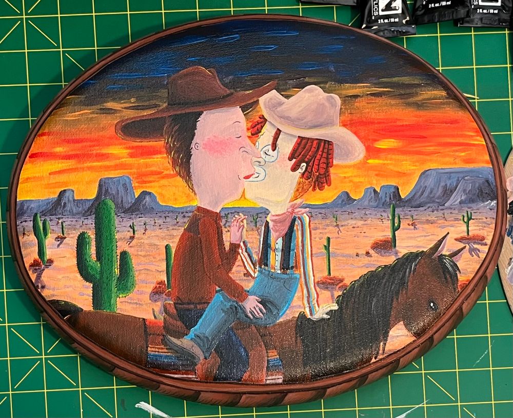 An acrylic painting on an oval canvas of Beavis and Butthead kissing while on horseback with a desert sunset and cacti in the background. Butthead is dressed as a cowboy with a red long sleeve and dark denim jeabs, so he’s riding the horse facing forward. Beavis is a rodeo clown, and he’s riding the horse backwards so he can kiss Butthead. Beavis has clown makeup on, and is wearing a terrible read dreadlock wig. His shirt is striped red, blue, yellow, and white. He’s wearing suspenders that hold up oversized light blue jeans. You can see his regular hair underneath. The whole thing looks magical. The shot is framed by a brown hand painted frame because the artist is a fucking idiot