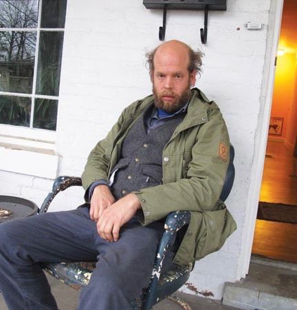 Will Oldham sitting in a metal chair on a porch with green parka on looking very Will Oldham.