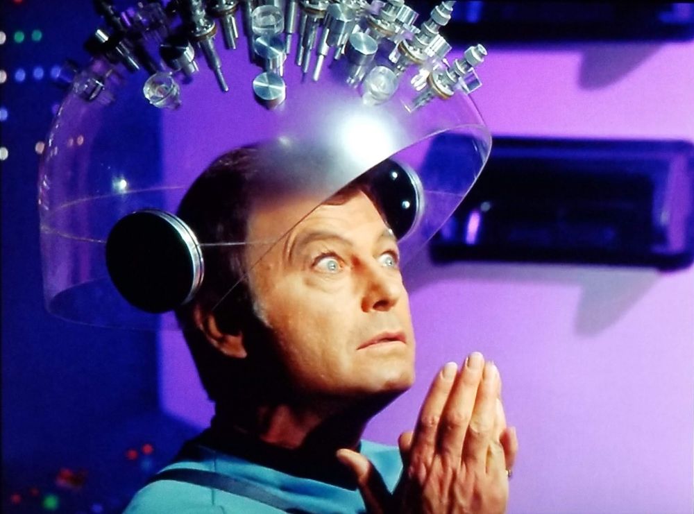 McCoy from TOS his hands in a prayer pose with a large clear device on his head