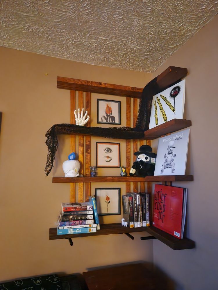 A handmade 4-tiered corner shelf with some small pieces of wood panel backing that frame three square prints. The shelf is cluttered with some Halloween decor, library books, and several vinyl records. 