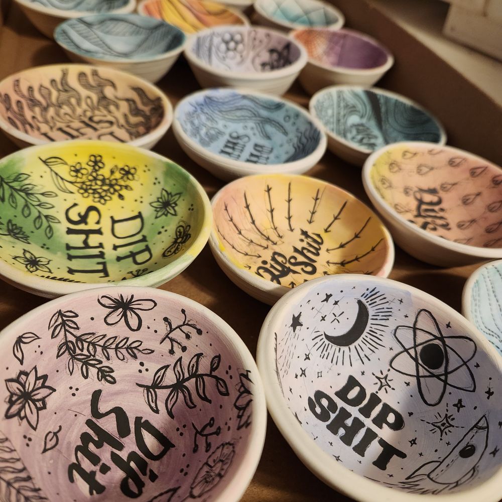 Several bisqued and underglaze painted tiny dishes, all with various designs and colours, plus the text "dip shit"