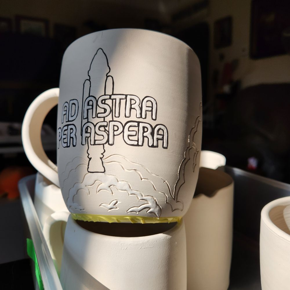 Partially decorated bisque mug with the text 'Ad aspera per aspera' overlaying the outline of a rocket. mug is sitting on a stack of other ceramic mugs that are uncompleted in broken sun beam