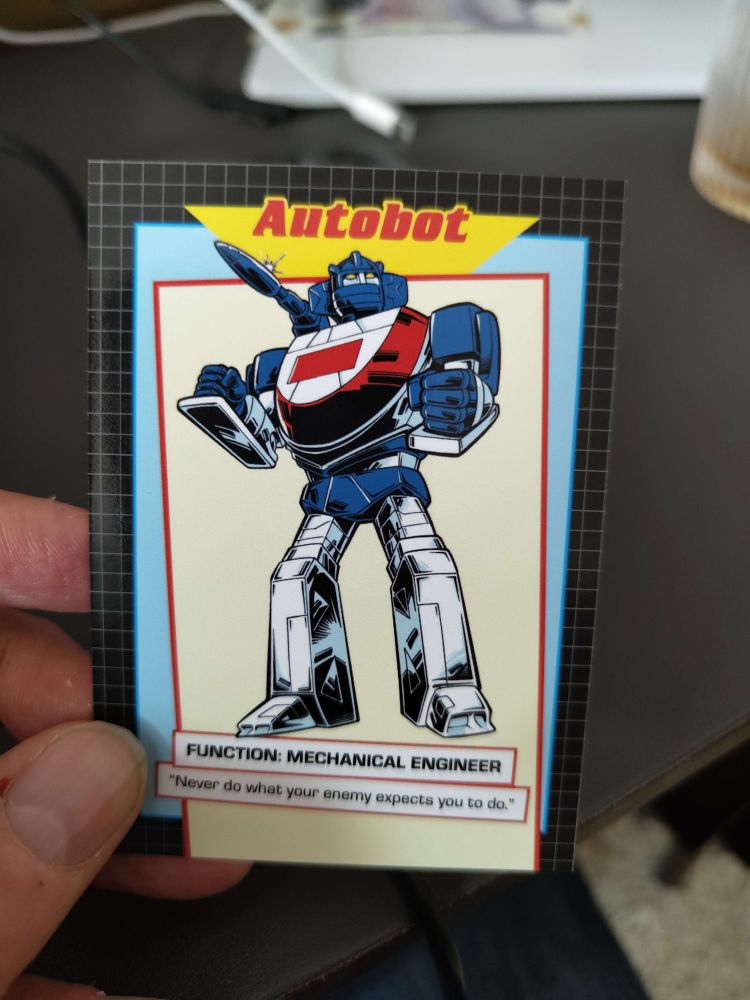A hand holding up a trading card featuring a white and blue robot. The title says "Autobot" and the bottom says "Function: Mechanical Engineer." It has the quote "Never do what your enemy expect you to do."