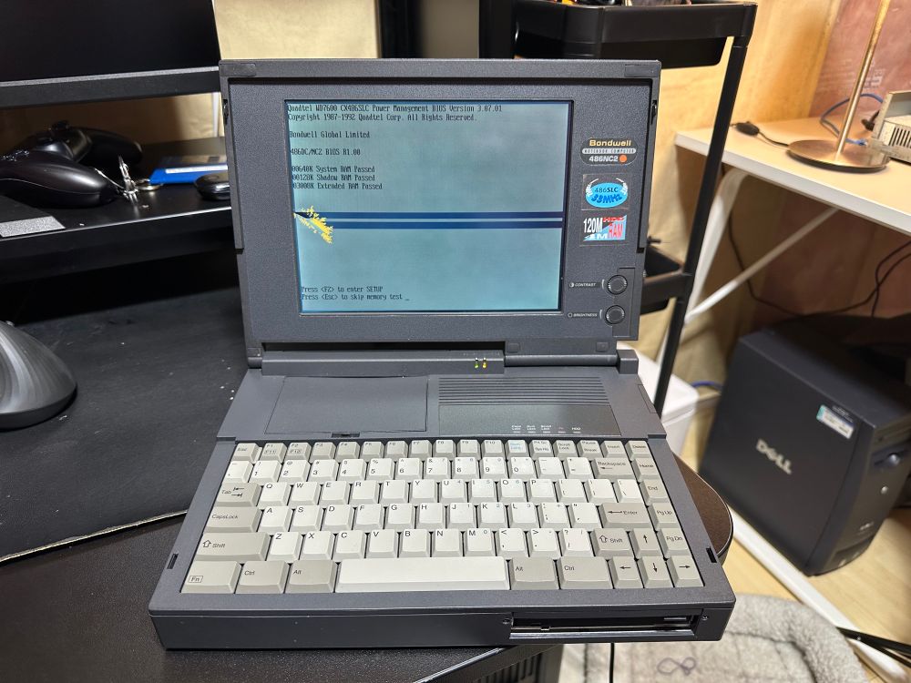 On a desk in a brightly lit basement, a very old laptop sits on a desk. It’s dark grey with a light grey keyboard. There is an early 2000s Dell tower on the floor in the background. The screen of the old laptop is black and white with a washed out image, as it’s a passive matrix screen. There is a spot of yellow-looking pixels on the left of the screen roughly in the middle, that appears to be gradually spreading. It’s causing two large horizontal lines to spread across the screen. This is gradual and irreversible degradation of the screen.
