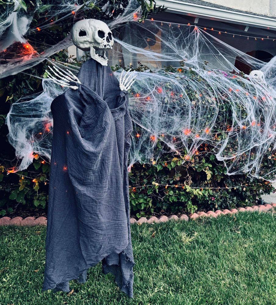 A homemade ghoul with skeletal jazz hands hangs from a tangerine tree in a suburban front yard. The ghoul’s skull head is laughing as their gray robe hangs in tatters. There are orange lights and spiderwebs on the tree and bushes in the background. 