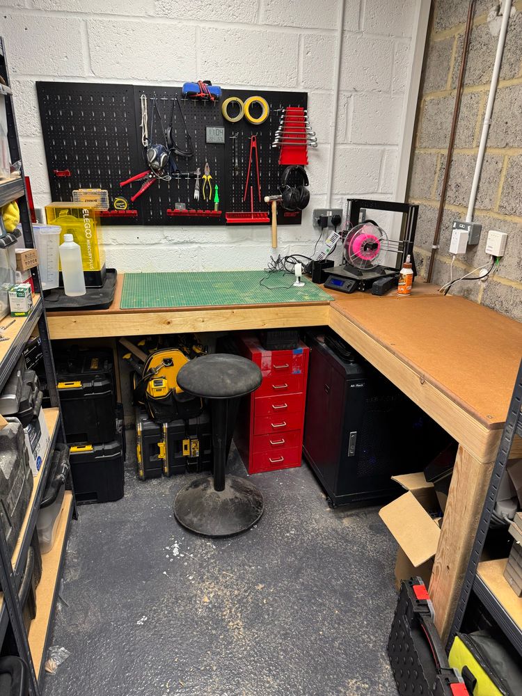 Photo shows the workshop I’m building in my garage. There’s an L shaped bench across the back wall with the return coming along the right wall. There’s pegboard on the wall with tools hung on and a 3D printer in the corner. 