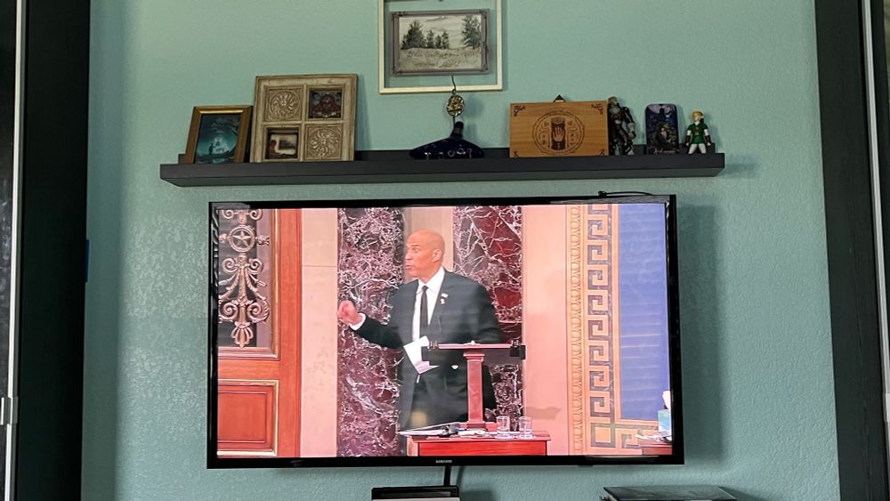 Snapshot of a wall mounted tv showing Senator Booker after breaking Thurmond’s record. The shelf above has assorted small Zelda art and a watercolor of trees. Library books are stacked below to the right and a tray of dishes left. 