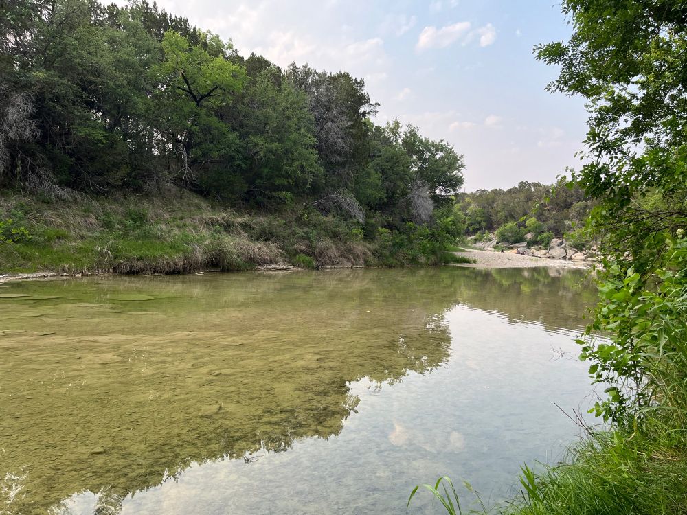 View from the shore across a bend in a shallow river with a limestone bed and vegetation growing near the water. Faint depressions of fossilized tracks are visible in the clear water. The vegetation is typical of post-oak savannah. 