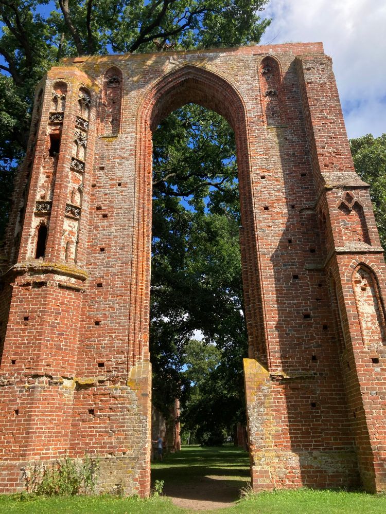 Reste eines Fensterbogens einer Ruine aus Backstein. Im Hintergrund ist ein großer Baum zu sehen.