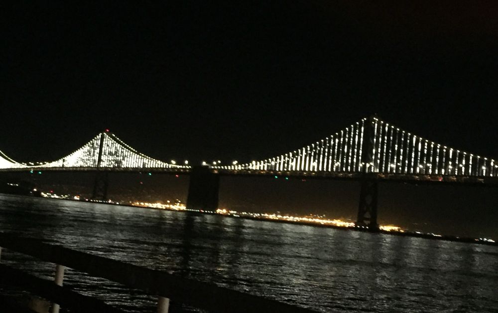 Blick auf die Bay Bridge bei Nacht. Die Brücke leuchtet durch eine weiße Lichtinstallation, die auch das Wasser erhellt während der Himmel schwarz ist.