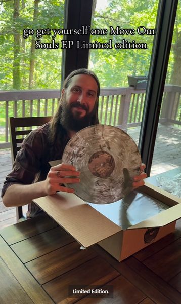 Avi Kaplan (wearing a brown shirt with the sleeves rolled up above his elbows) holds up the new vinyl record for Move Our Souls EP Limited Edition now on sale.  It has a lot of tans and browns on a white background of what looks like someone in the woods.  It looks like someone took the smear tool in Photoshop around the record to give a more fantasy-like quality to the picture.  This image is printed on both sides of the record.