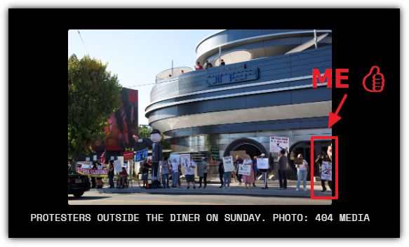 Protesters holding signs gather outside a diner, with a modern building and trees in the background.
