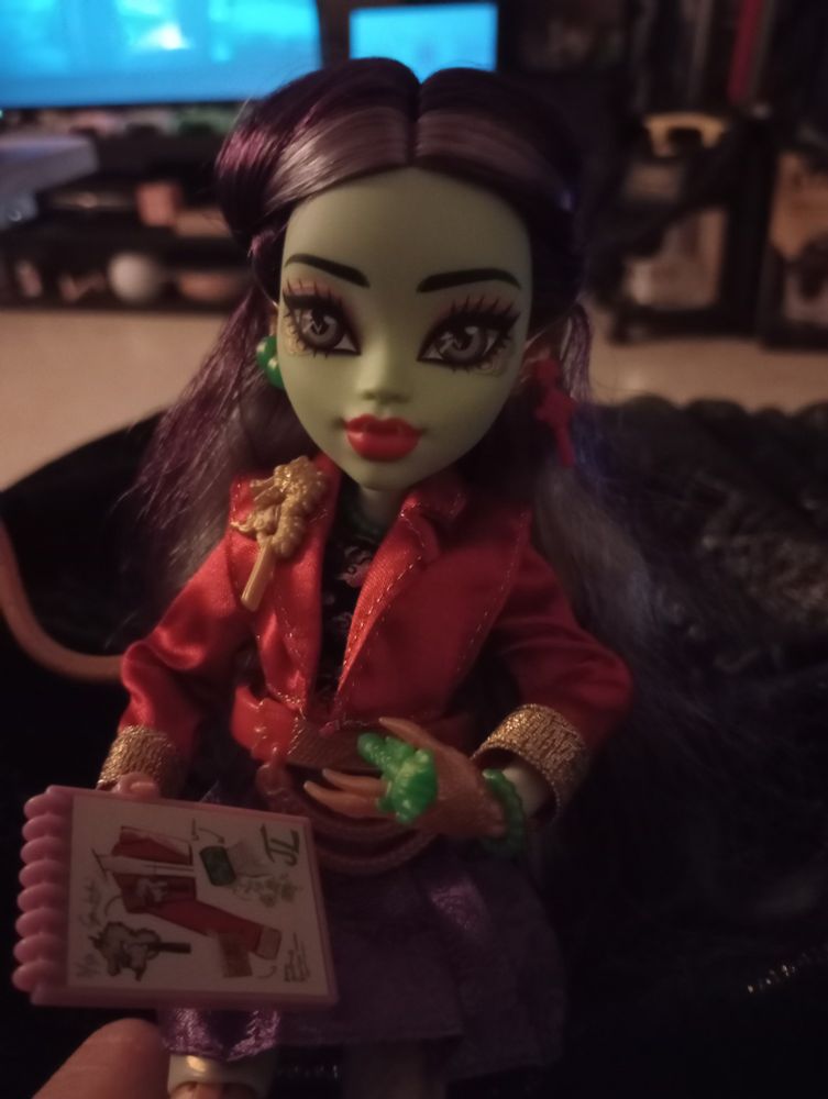 Photo of G3 Monster High doll Jinafire Long. She has a green skin tone (darker than Ghoulia's), a long golden dragon tail with jade fin, and her hands and legs have golden scales. 

She has a purple and black top, purple skirt, and red jacket. She also has quite a few jade colored accessories and some gold highlighting in her outfit.

Her hair is a dark purple with some lighter purple streaks. Her irises are brown with an inner circle of golden and jade. I have her holding a fashion design notebook and jade pen.