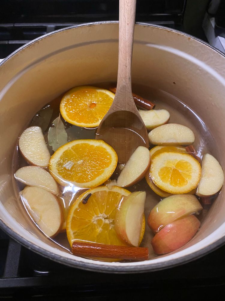 A white enamel Dutch oven with orange and apples slices, cinnamon sticks, cloves, bay leaves, and vanilla mixed with water. 