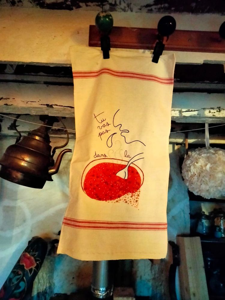 Torchon blanc rayé de rouge en haut et en bas. Il est brodé de l’expression : tu vas pas cracher dans la soupe. 
Un profil, une goutte qui tombe vers la soupe. La soupe est brodée de perles et de tubes dan les tons orange et doré sur une application de tissu orange. 
La broderie de perles débordé un peu sur le blanc du torchon. 

Le torchon est suspendu par des pinces sur une vieille poutre chaulée de blanc. Une vielle théière en cuivre lui tient compagnie. 
