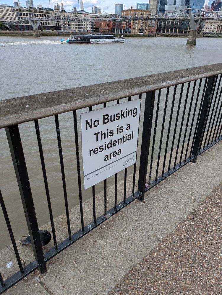Sign on a fence near a river reading "No Busking: This a residential area"